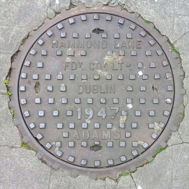 Ein metallener Gullideckel auf einer Straße in Dublin, Irland, mit eingravierter Schrift.