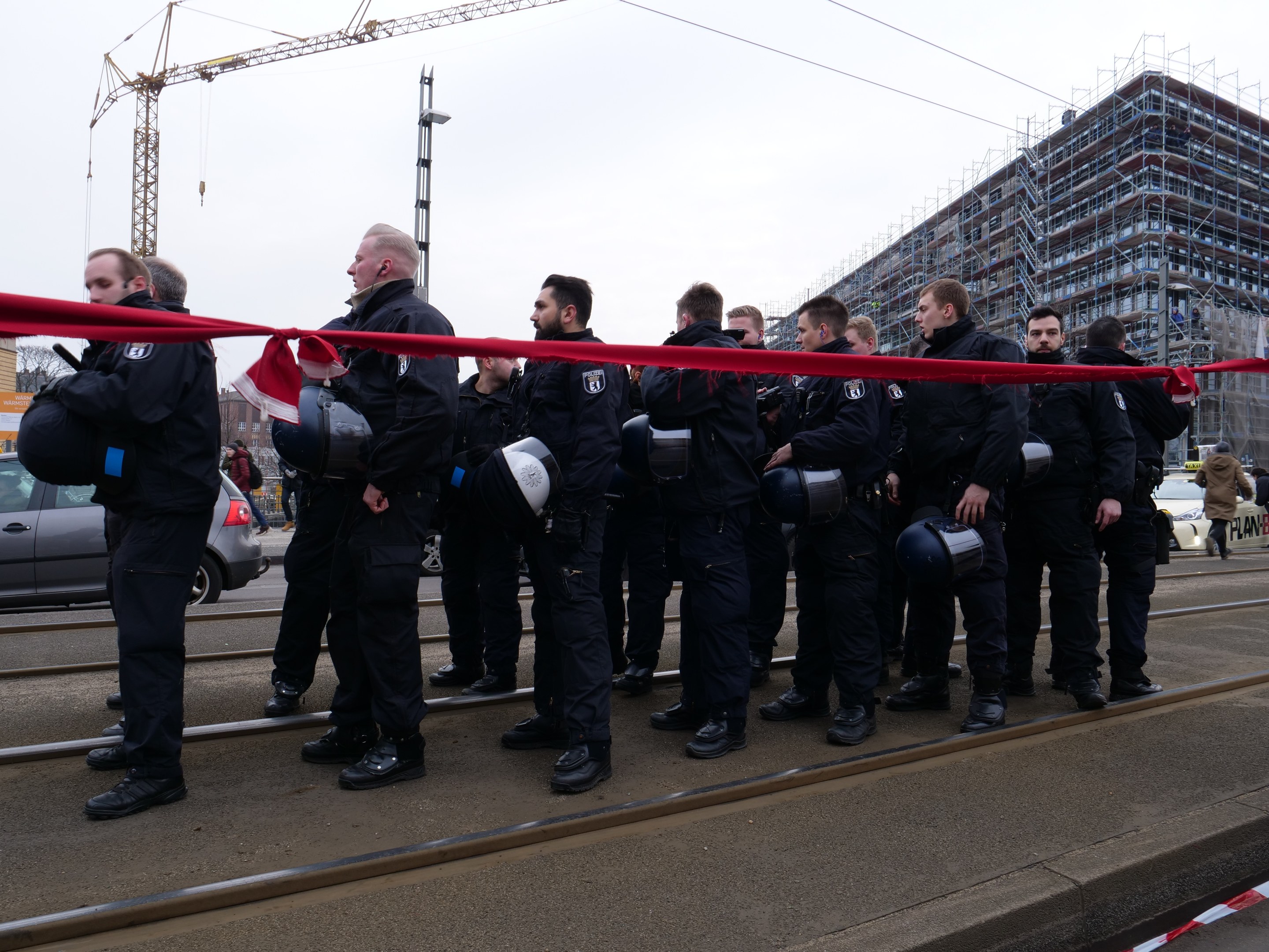 Gruppe von Polizisten in schwarzen Uniformen und Helmen auf einem Bahngeleis mit einer roten Schleife, Fahrzeuge und Passanten im Hintergrund unter einem klaren blauen Himmel.