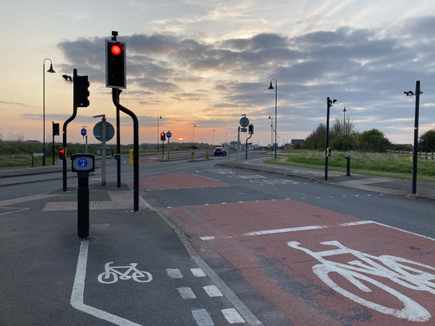 Eine Radspur mit einer roten Ampel, Fahrzeuge auf der Straße, Laternenpfähle, Schilder, Gras, Bäume und ein bewölkter Himmel im Hintergrund.
