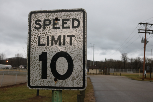 Geschwindigkeitsbegrenzungsschild an der Seite einer Straße mit umgebenem Gras, Strommasten, einem Zaun, Häusern, Bäumen und einem bewölkten Himmel.