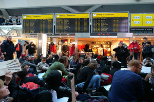 Große Gruppe von Menschen in einem Flughafen, einige mit Taschen und Papieren sitzend, andere stehend, mit Informationsschildern, Schaufensterpuppen in Kleidern und Deckenbeleuchtung, was auf eine Protestaktion hindeutet.