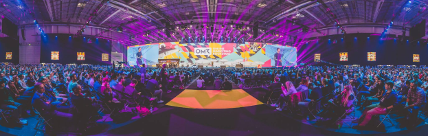 Eine große Menschenmenge, die auf Stühlen sitzt und einer hell erleuchteten Bühne mit mehreren stehenden Personen gegenüber sitzt, wahrscheinlich bei einem Online-Marketing-Rockstars-Event, umgeben von Bildschirmen.