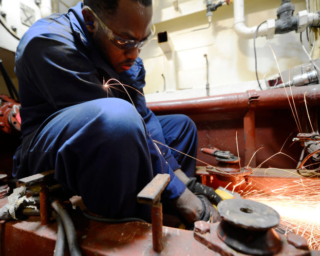 Ein Schweißer mit Brille arbeitet an einem Metallstück und erzeugt Funken in einer Fabrik mit Rohren und anderen Gegenständen im Hintergrund.