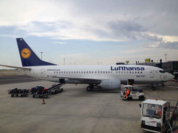 Ein Lufthansa Airbus A320-200 steht am Frankfurter Flughafen, umgeben von verschiedenen Bodenfahrzeugen, Wagen, Absperrgittern, Pfosten und Texttafeln unter einem bewölkten Himmel.