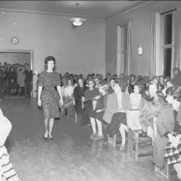Eine Frau in einem schwarzen und weißen Kleid läuft über den Laufsteg während einer Modenschau für Frauenrechte, mit einer Menge, die auf Stühlen sitzt und einer Uhr und Lampen an der umgebenden Wand.