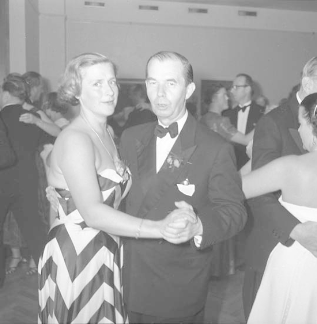 Ein Mann in einem schwarzen Blazer, weißem Hemd und schwarzen Hosen tanzt mit einer Frau in einem unbekannten Outfit auf einer formellen Veranstaltung, indem sie sich an den Händen halten und zur Musik wiegen, während sie in der Nähe einer Wand tanzen.