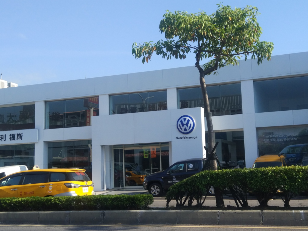 Volkswagen-Händler mit Glaswänden, parkenden Autos, umgeben von Pflanzen und einem Baum, mit Himmel im Hintergrund.
