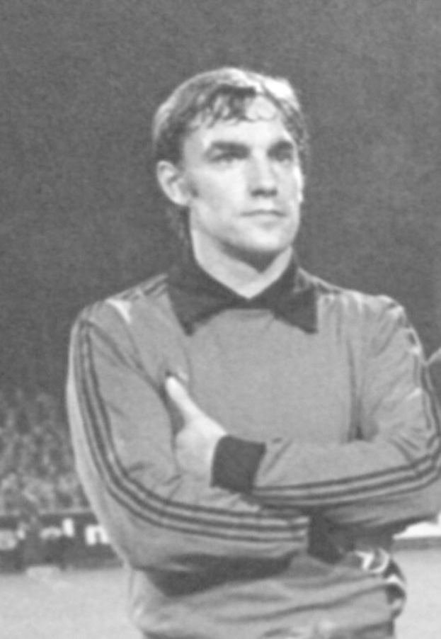 Schwarzes und weißes Foto eines Mannes, der mit verschränkten Armen auf einem Fußballfeld steht und von einer Gruppe sitzender Menschen im Hintergrund umgeben ist.