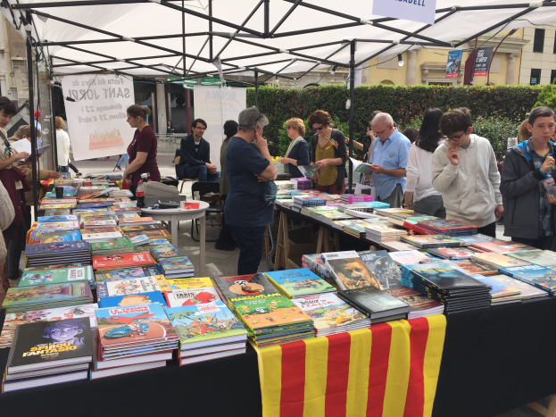 Eine Gruppe von Menschen auf einer Buchmesse in Barcelona, die um einen mit einem Tuch bedeckten Tisch stehen, der mit Büchern gefüllt ist, einige halten Bücher in den Händen, mit einem Banner mit Text, Pflanzen und Gebäuden mit Fenstern im Hintergrund unter einem klaren blauen Himmel.