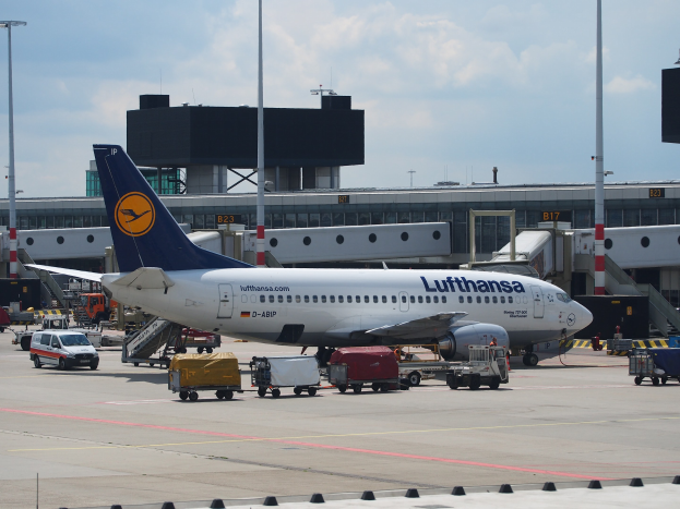 Lufthansa Airbus A320-200 am Frankfurter Flughafen mit Bodenfahrzeugen und Wagen vorne, einem Gebäude und Laternen im Hintergrund vor einem klaren blauen Himmel.