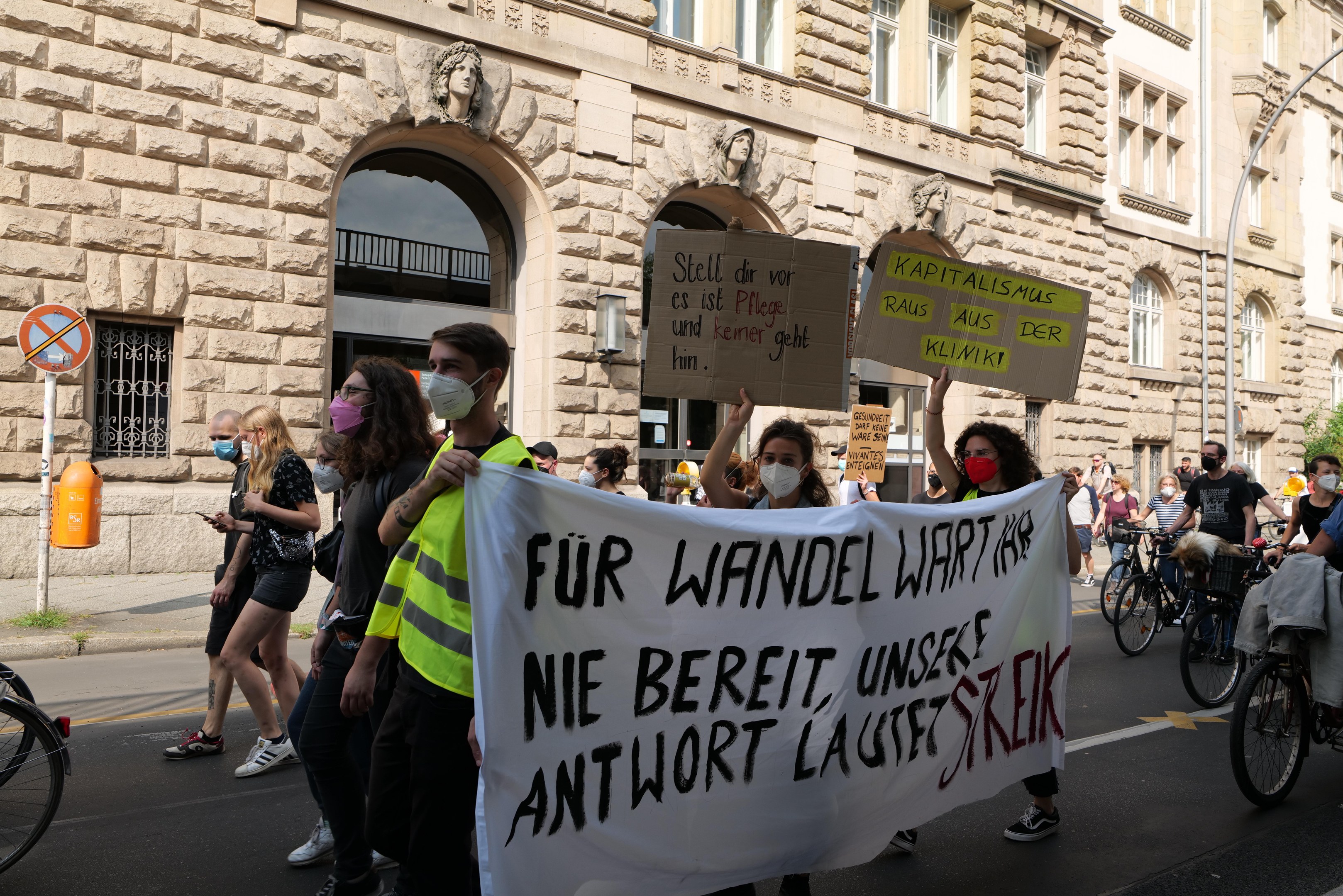 Eine Gruppe von Menschen marschiert bei einer Protestaktion gegen die Anti-Kriegs-Bewegung in Berlin, einige halten Schilder und andere fahren Fahrräder, im Hintergrund ein Gebäude mit Fenstern, Bögen, Säulen und Skulpturen.