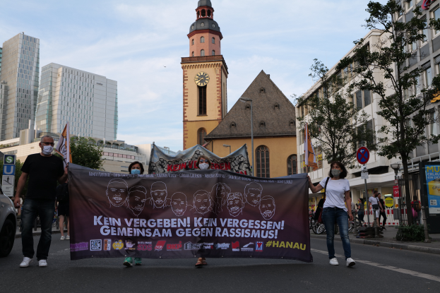 Eine Gruppe maskierter Personen marschiert auf einer Straße und hält ein Transparent, mit einem geparkten Auto auf der linken Seite, Gebäuden, Bäumen, Schildern, Polen und einem Kirchturm im Hintergrund unter einem klaren blauen Himmel.