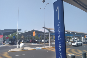 Eine Mautstelle am Flughafen mit parkenden Autos davor, beleuchtet von Straßenlaternen, mit Schildern, Pfählen, einem Schuppen, Bäumen und einem klaren blauen Himmel im Hintergrund.