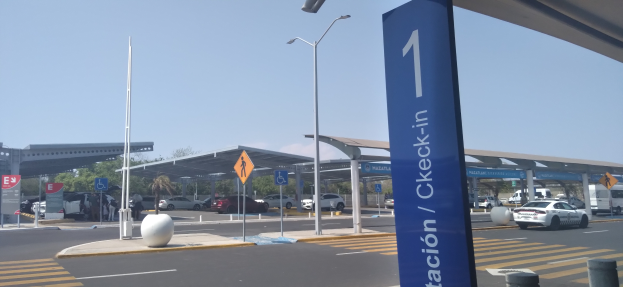 Eine Mautstelle am Flughafen mit parkenden Autos davor, beleuchtet von Straßenlaternen, mit Schildern, Pfählen, einem Schuppen, Bäumen und einem klaren blauen Himmel im Hintergrund.