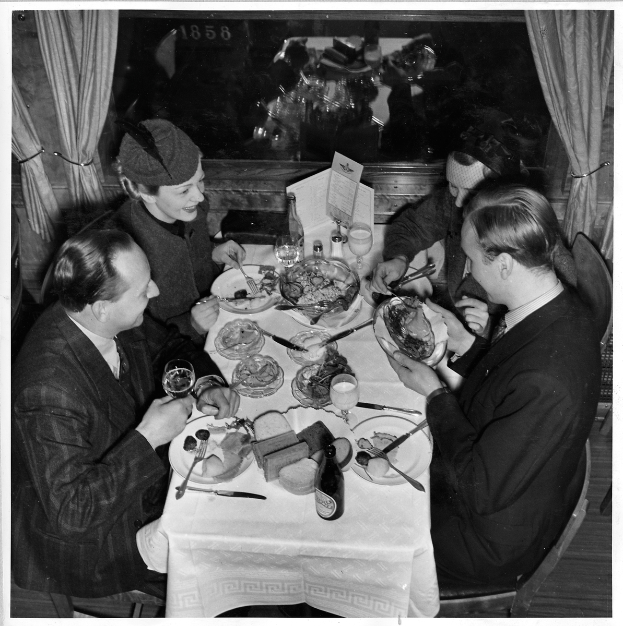 Eine Gruppe von Menschen sitzt um einen Tisch mit Tellern, Gläsern, Flaschen und Besteck, mit Vorhängen im Hintergrund.