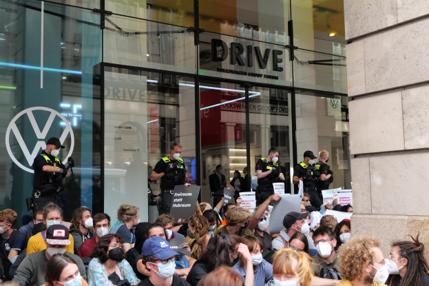 Große Gruppe von Menschen mit Masken und Schildern vor einem Glaswandgebäude, einige tragen Mützen und halten Kameras, was auf eine Demonstration in Melbourne, Australien, hindeutet.
