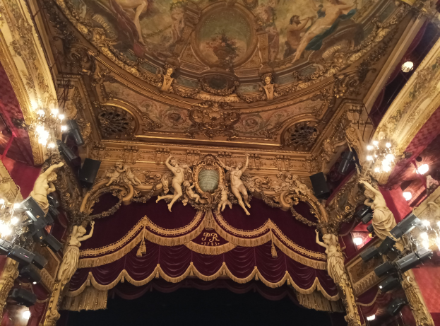 Das Innere eines Theaters mit einem roten Vorhang, Kronleuchtern, Skulpturen, Lichtern, einer Deckenmalerei und einer Bühne mit ein paar Menschen unten.