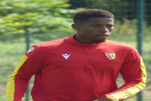 Junger Mann in rotem und gelbem Fußballtrikot auf einem Feld mit einem Zaun und Bäumen dahinter, der konzentriert als Flügelspieler erscheint.