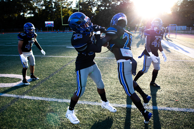 Junge Männer in Helmen und Schuhen spielen Fußball auf einem von Bäumen umgebenen Feld unter einem klaren blauen Himmel.