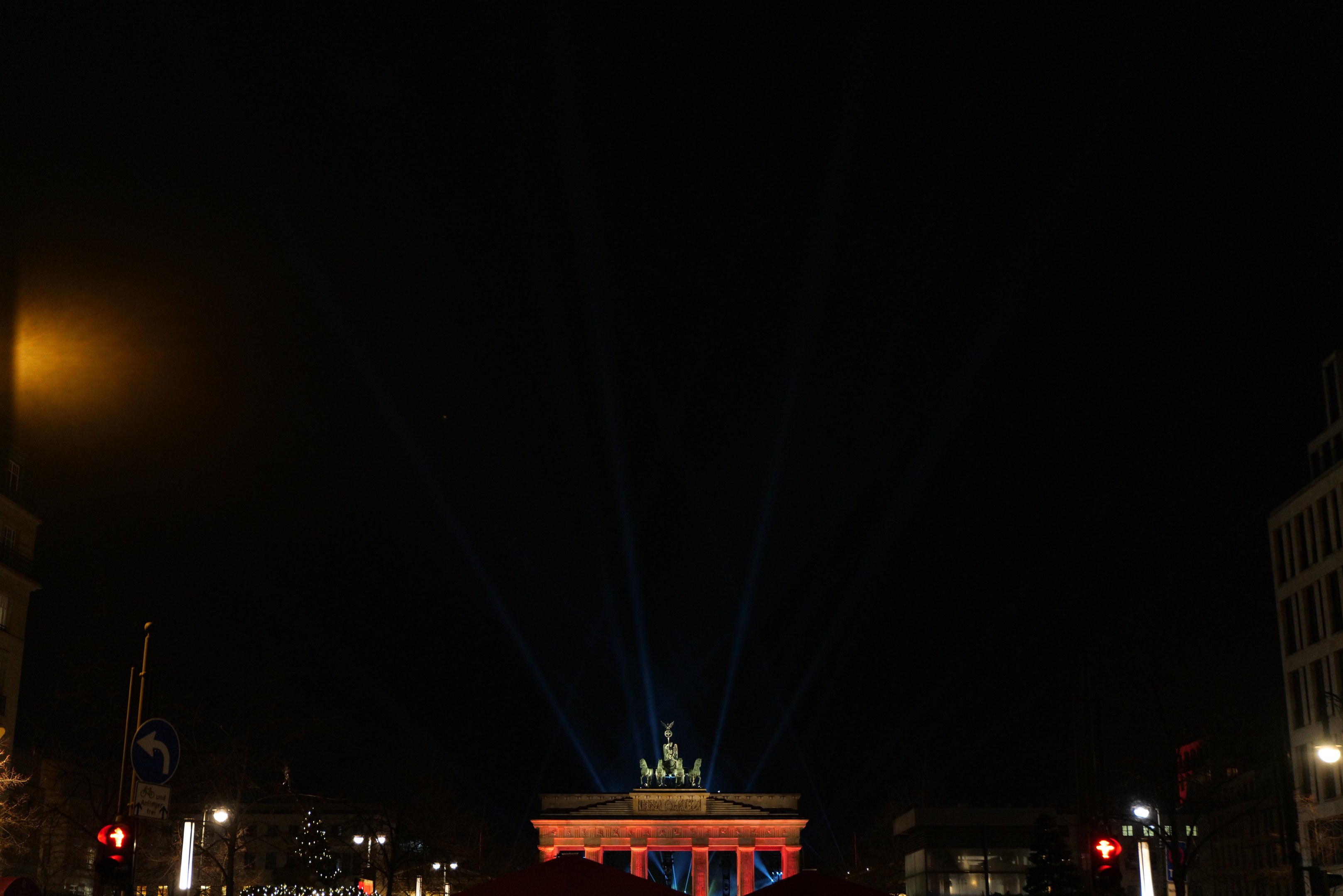 Das Brandenburger Tor in Berlin nachts, beleuchtet von Straßenlaternen, mit umliegenden Gebäuden, Bäumen, Pfählen, Verkehrszeichen, Schildern und Zelten, vor einem dunklen Himmel.