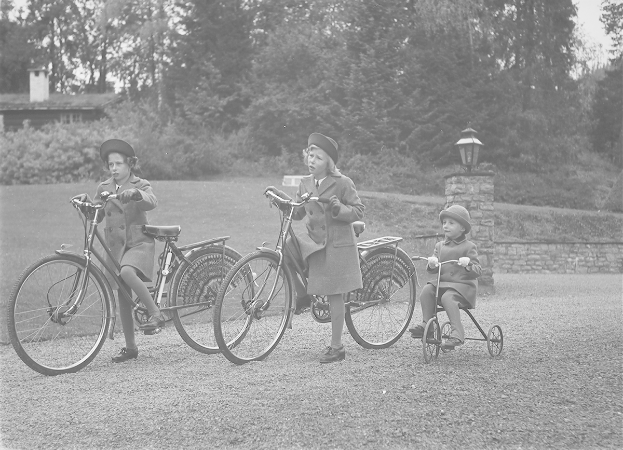 Drei Kinder fahren Fahrräder in einem Park, tragen Mäntel und Mützen, wobei eines auf einem Dreirad sitzt, vor einem Hintergrund aus Bäumen, Pflanzen, einem Haus und einem Laternenpfahl, in Schwarz-Weiß.