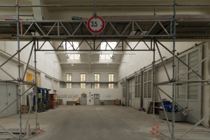 Großer Lagerraum mit ausgedehnter Gerüstkonstruktion, Verkehrsleitkegeln, Metallstangen, einem Schild, Maschinen, einer Tür, Fenstern, Wänden, Deckenleuchten und einem Dach.
