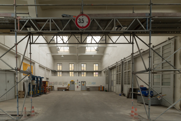 Großer Lagerraum mit ausgedehnter Gerüstkonstruktion, Verkehrsleitkegeln, Metallstangen, einem Schild, Maschinen, einer Tür, Fenstern, Wänden, Deckenleuchten und einem Dach.