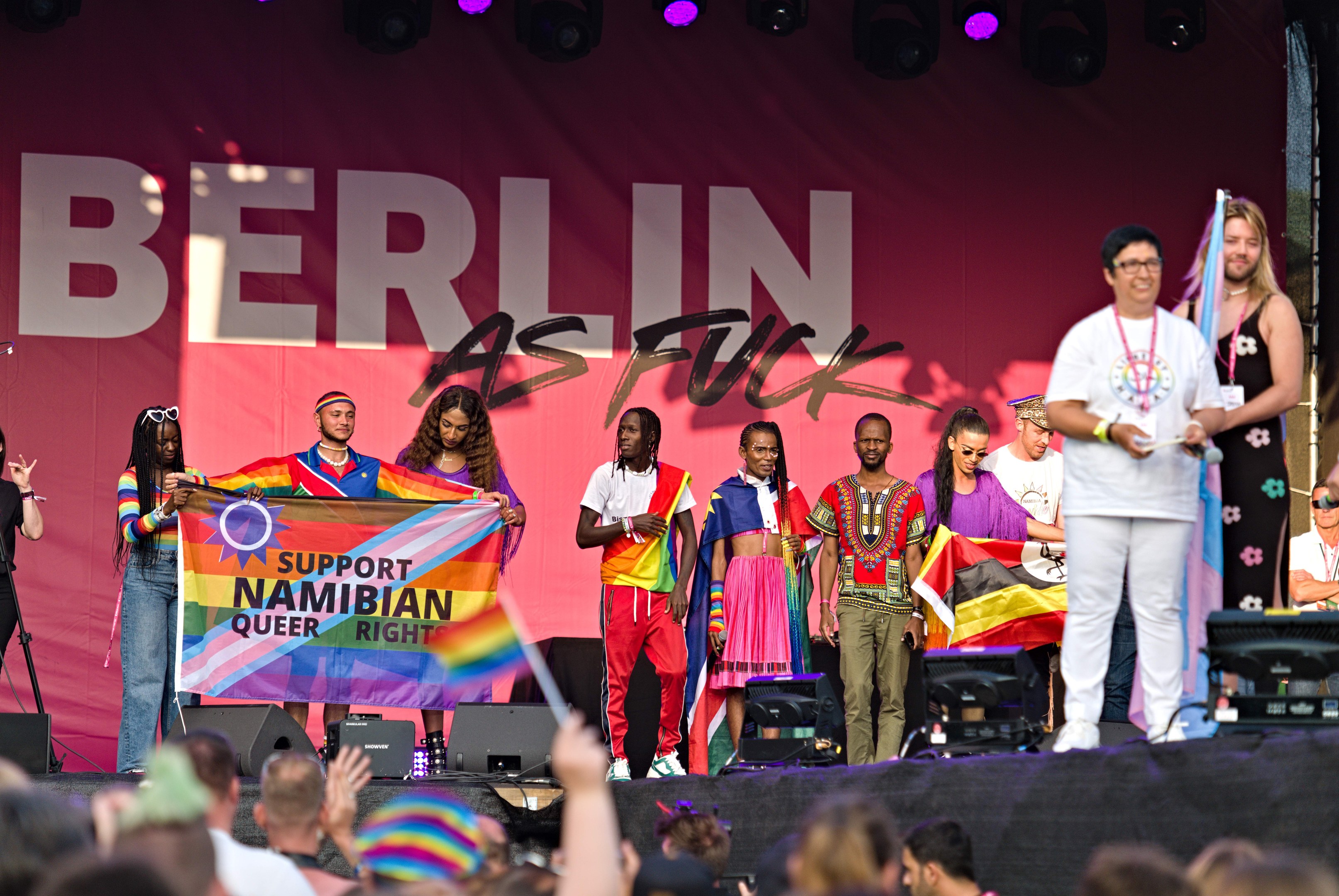 Gruppe von Menschen auf einer Bühne mit einem "Berlin as Fuck"-Schild, Lautsprechern, Mikrofonen und einer Menge unten mit hochgereckten Händen.