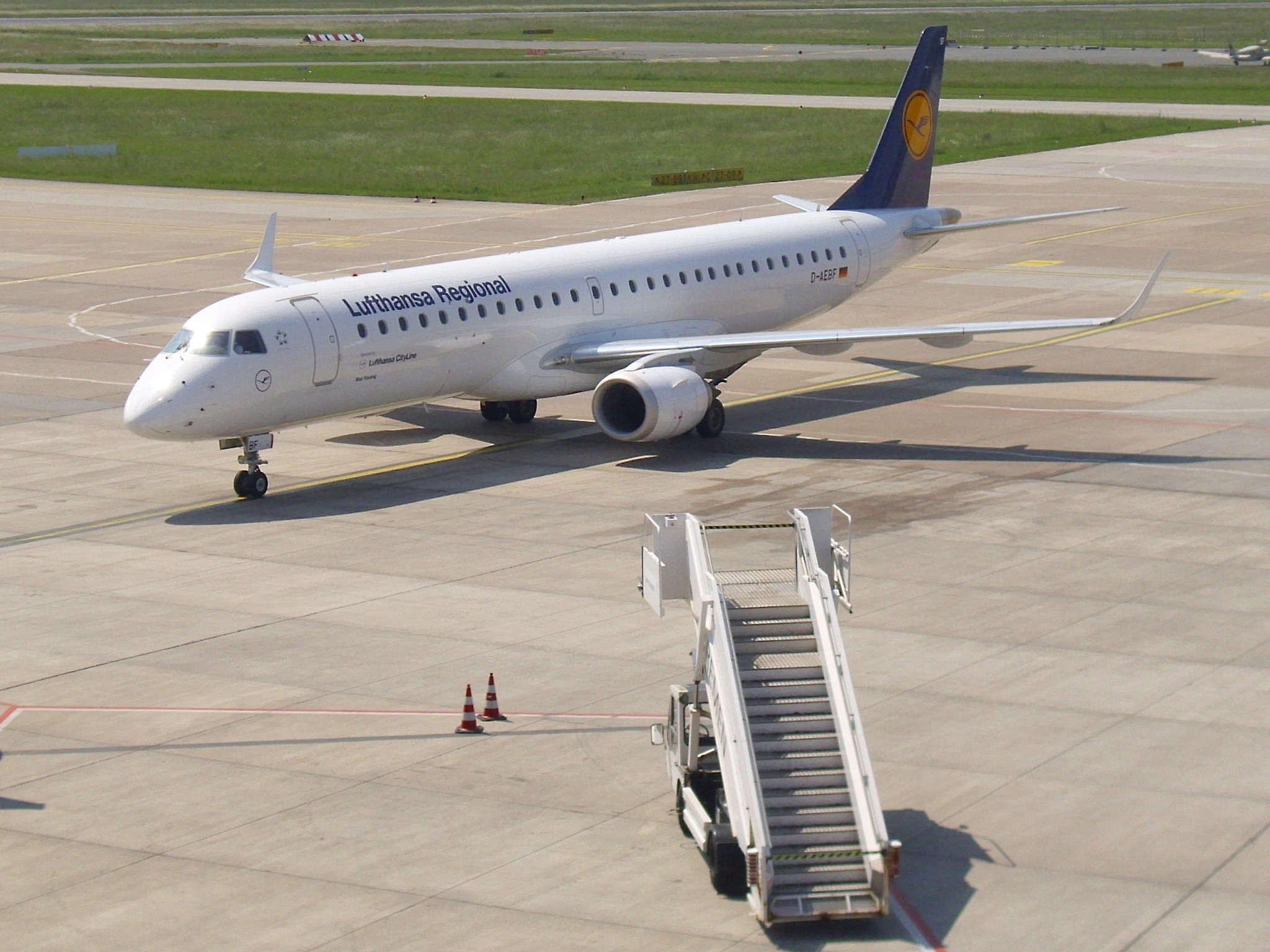 Lufthansa Regional Airbus A320-200 auf der Rollbahn am Frankfurter Flughafen, mit Verkehrskegeln und einem Treppefahrzeug in der Nähe, vor grünem Gras, Bäumen und einem klaren blauen Himmel.