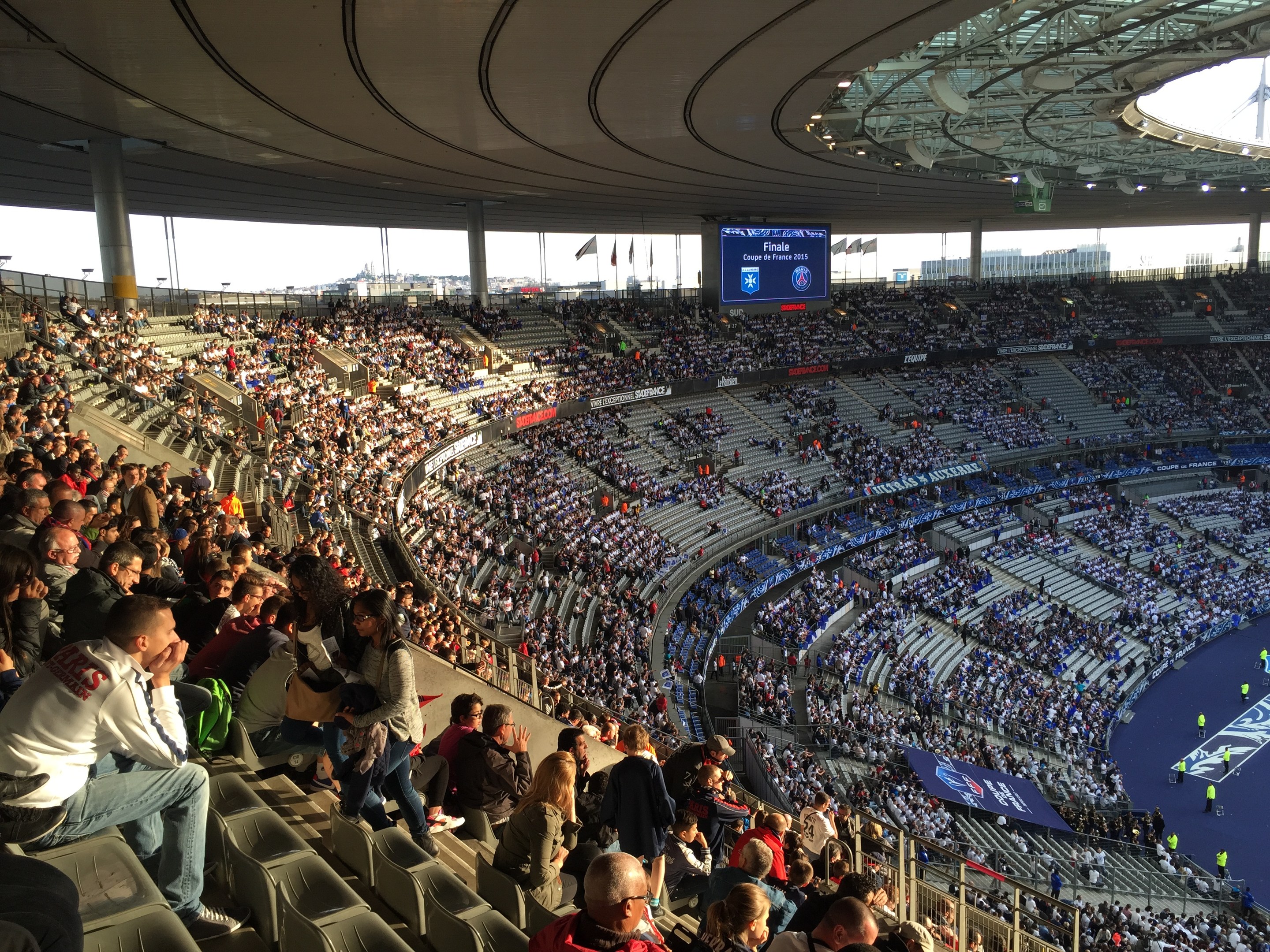 Großes Publikum in einem Stadion, das ein Fötballspiel verfolgt, mit einer Bühne rechts, Fahnen, Masten, einem Bildschirm und der Allianz Arena in München im Hintergrund.