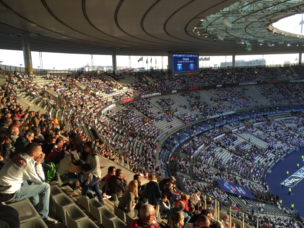 Großes Publikum in einem Stadion, das ein Fötballspiel verfolgt, mit einer Bühne rechts, Fahnen, Masten, einem Bildschirm und der Allianz Arena in München im Hintergrund.