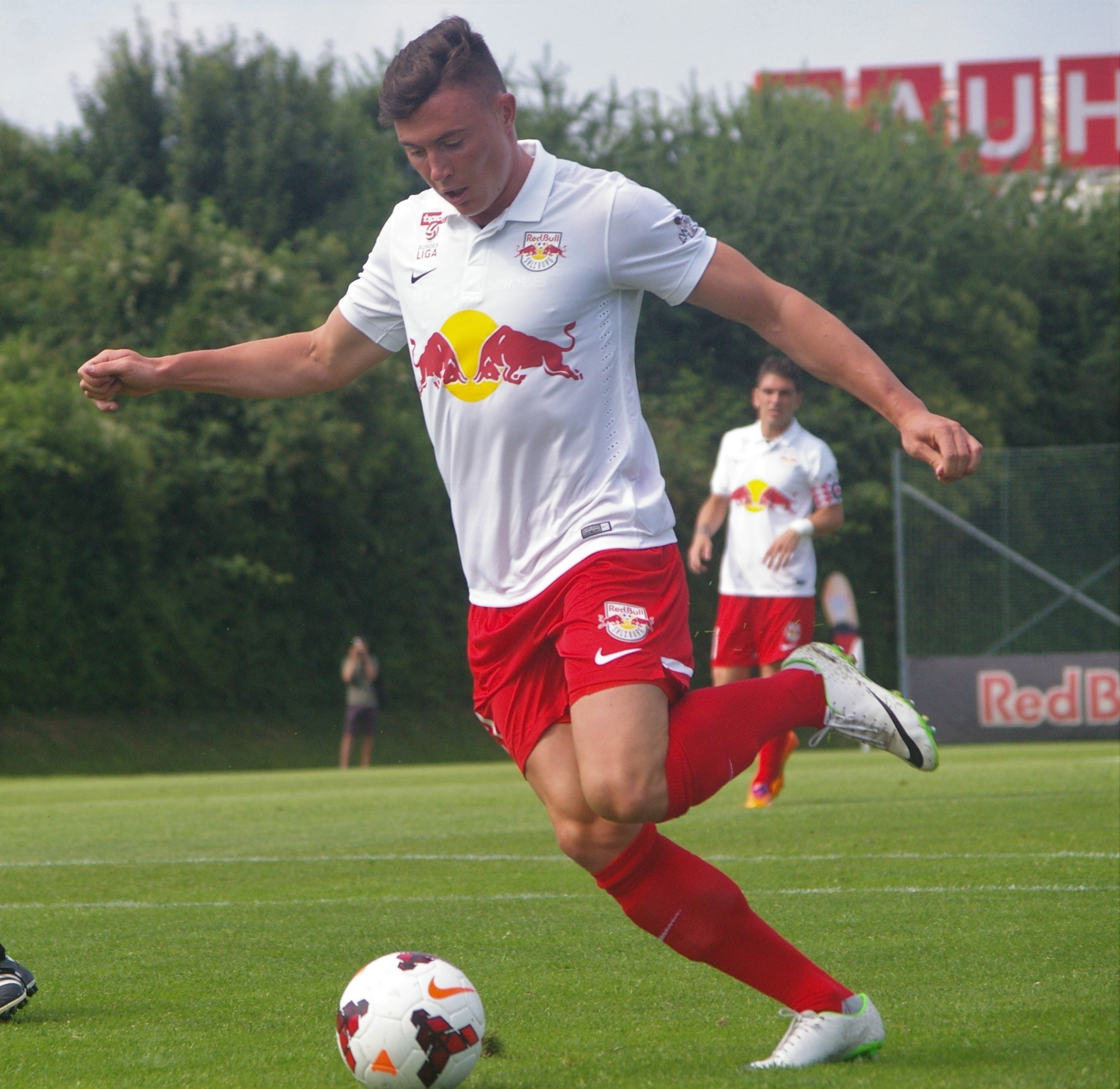 Ein professioneller Fuballspieler in einem roten Trikot schießt einen Ball auf einem grünen Feld mit Bäumen und einem klaren blauen Himmel im Hintergrund, in der Nähe einer Tafel mit der Aufschrift "RB Leipzig."