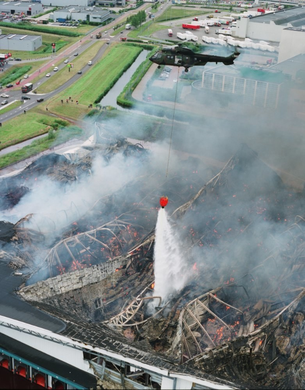 Ein Hubschrauber fliegt über ein großes, von Feuer zerstörtes Gebäude mit Rauch, der daraus aufsteigt, vor einem Hintergrund aus städtischen Strukturen, Fahrzeugen auf der Straße und Bäumen, mit Text unten.
