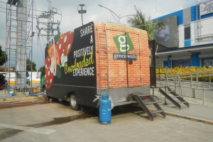 Ein weißer Lkw vor einem Gebäude mit einer Tafel, auf der "Teilen Sie eine positiv überladene Erfahrung" steht, mit Stufen, einem Zylinder am Boden, Geländern, Strommasten mit Kabeln, Laternen, Bäumen und einem bewölktem Himmel im Hintergrund.