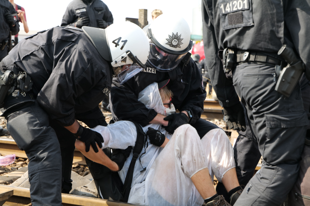 Eine Gruppe von Polizisten in schwarzer Einsatzkleidung verhaftet einen Mann in einem weißen Kleid auf den Schienen, mit anderen Menschen und dem Himmel im Hintergrund.
