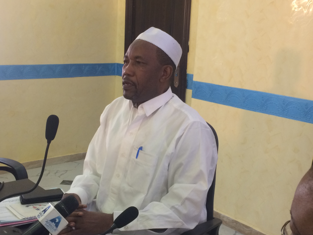 Ein Mann in einem weißen Gewand und einer Mütze sitzt an einem Tisch mit Mikrofonen, Papieren und anderen Gegenständen und spricht, während rechts von ihm eine Person mit Brille sitzt, in einem Raum mit einer Wand und einer Tür im Hintergrund.