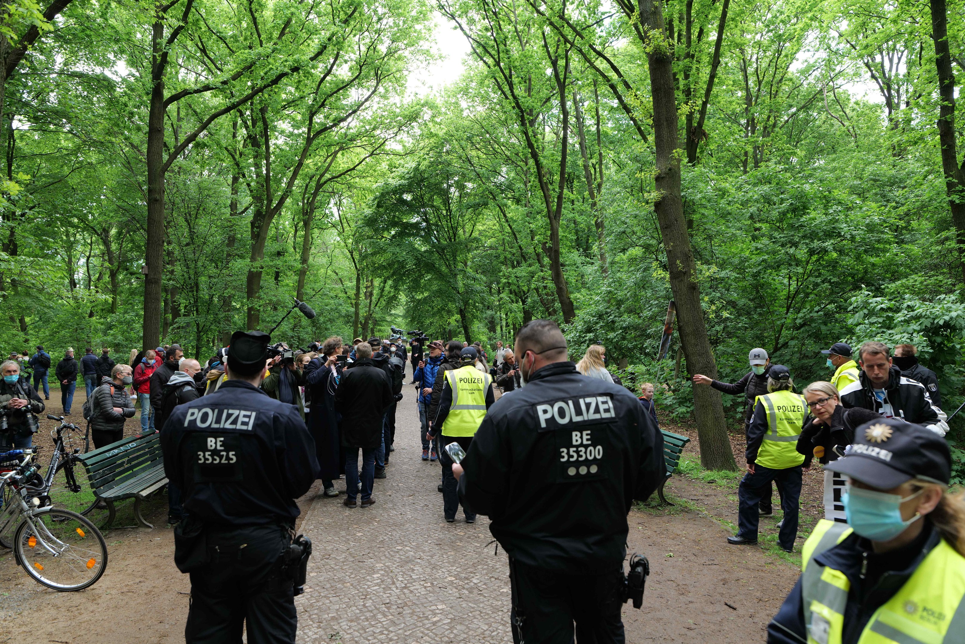 Polizisten vor einer Menge während einer Outdoor-Veranstaltung, mit Fahrrädern und einer Bank im Vordergrund und Bäumen im Hintergrund.