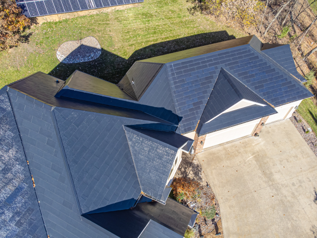 Ein Haus mit Solarmodulen auf dem Dach, umgeben von Bäumen, Gras und einem Zaun, aus der Vogelperspektive betrachtet.
