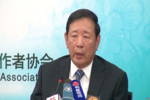 Ein Mann im Anzug sitzt an einem Tisch mit Mikrofonen und hält eine Pressekonferenz, mit einem Banner im Hintergrund.