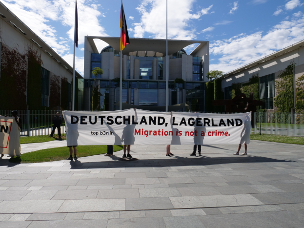 Gruppe von Menschen mit einer Tafel, auf der 'Deutschland, Lagerland Migration ist kein Verbrechen' steht, vor einem Gebäude mit Fenstern, umgeben von Gras, einem Metallzaun, Pflanzen, Bäumen und Fahnen mit Stangen unter einem bewölkten Himmel.