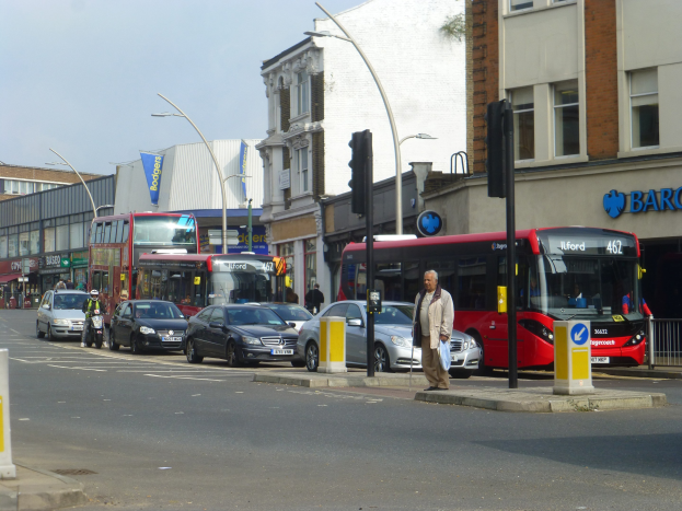 Ein roter Doppeldeckerbus fährt auf einer Stadtstraße mit hohen Gebäuden, Fußgängern, einem Radfahrer, Verkehrsampeln, Laternen und Schildern unter einem bewölkten Himmel.