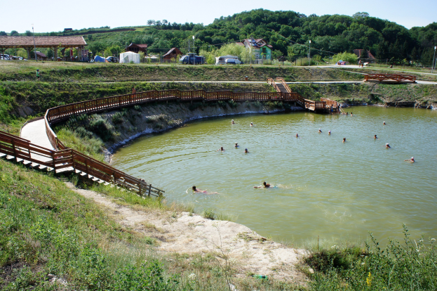 Gruppe von Menschen, die in einem Gewässer schwimmen, umgeben von üppiger grüner Vegetation, einer Brücke mit Geländer und Treppen, Hütten, Fahrzeugen, Pfählen und einem klaren blauen Himmel im Hintergrund.