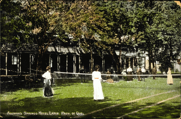 Ein altes Foto einer Gruppe, die auf einem Rasenplatz vor dem Abenakis Springs Hotel in Provo, Quebec, Tennis spielt. Einige halten Rackets, andere sitzen, ein Netz in der Mitte, Bäume und ein Gebäude im Hintergrund, sowie Text unten auf dem Bild.