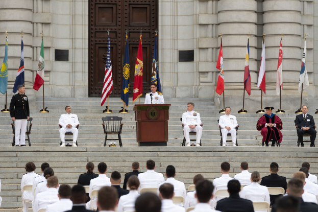 Gruppe von Menschen in weißen Uniformen auf den Stufen eines Gebäudes sitzend mit Flaggen zu beiden Seiten eines Podiums vor einem großen Gebäude mit einer Tür während einer feierlichen Zeremonie.