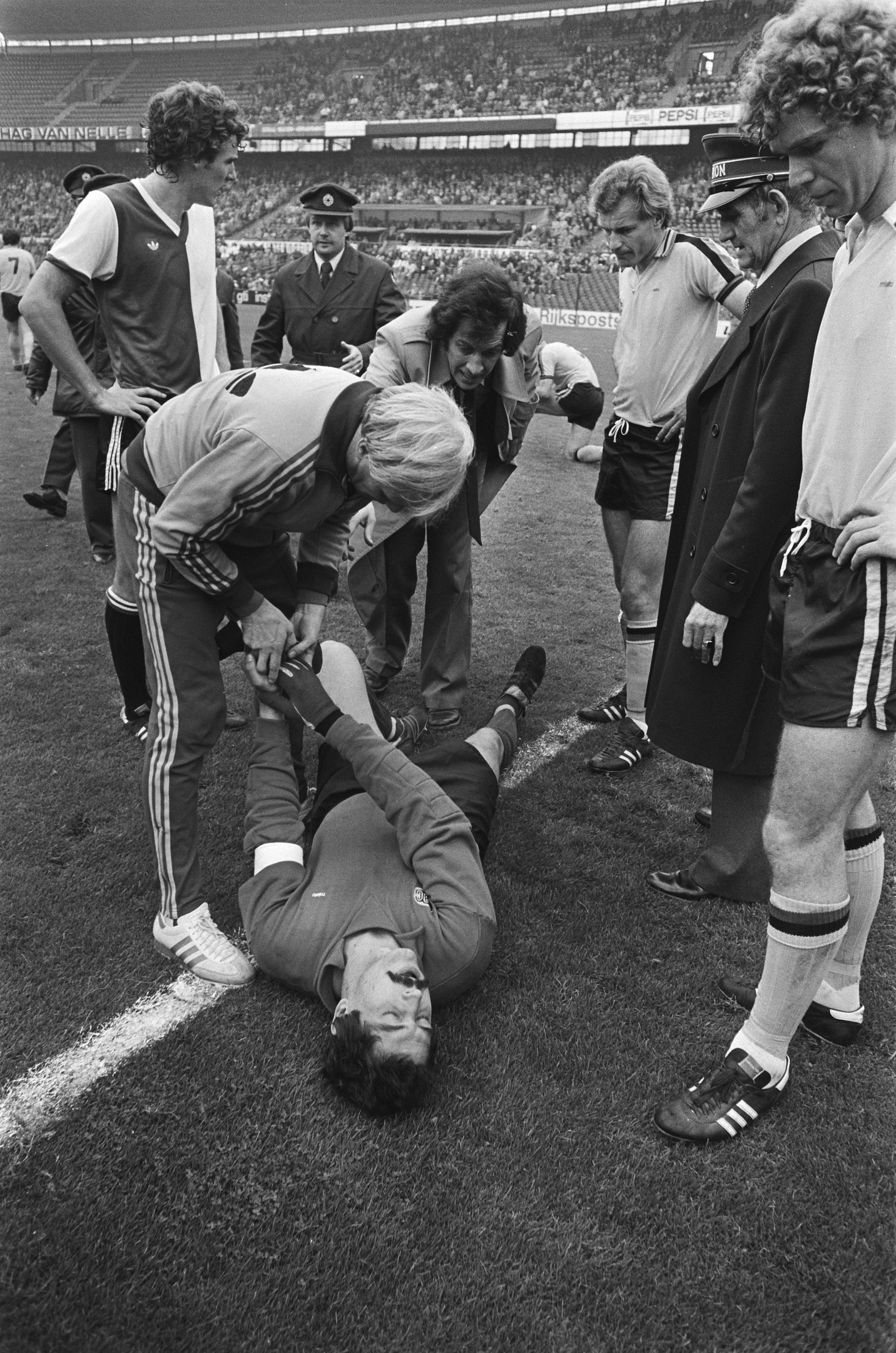 Schwarzes und weißes Bild eines Fußballspielers, der auf dem Boden liegt und von einer Gruppe von Männern umgeben ist, mit einer Menge im Hintergrund eines Stadions.