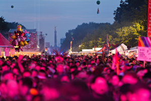 Eine große Menschenmenge auf einem nächtlichen Musikfestival mit bunten Lichtern, Ballons, Zelten, Bannern, Fahnen, Bäumen, einem Turm und einem sternenklaren Himmel im Hintergrund.