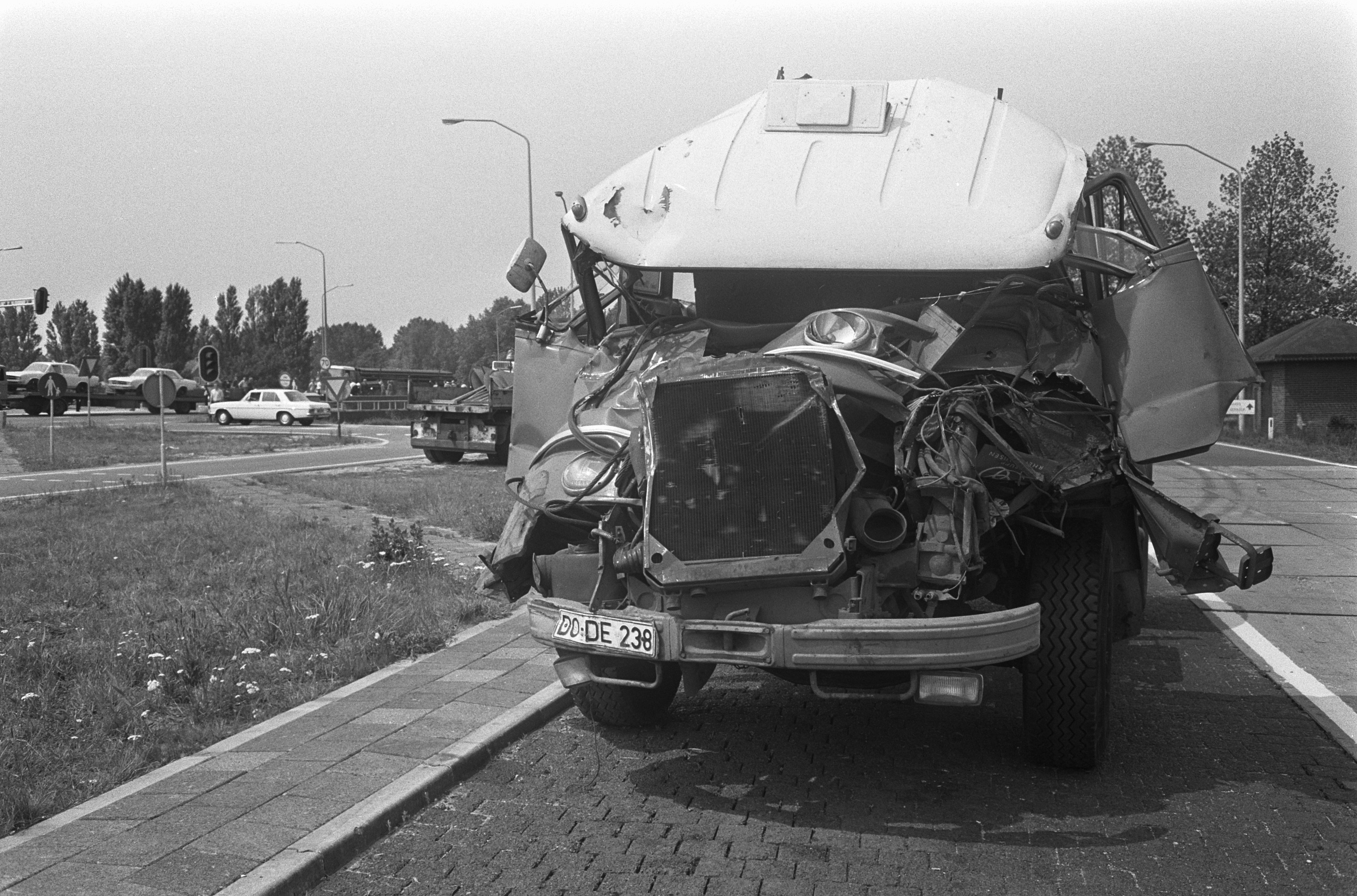 Ein Schwarz-Weiß-Foto eines schwer beschädigten Autos am Straßenrand mit eingedellter Motorhaube und zersplitterten Fenstern, mit anderen Fahrzeugen, Laternenpfählen, Bäumen und einem Haus im Hintergrund.