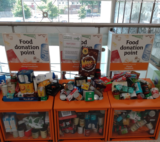 Orange Schränke voller Lebensmittelspenden, darunter Flaschen, Dosen und Kartons, mit Informationsschildern in der Nähe und einem Blick durch Glasfenster auf Fahrzeuge, Bäume, Gebäude und Himmel.