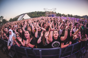 Menschenmenge auf einem Musikfestival mit erhobenen Händen vor einem Geländer, Bäume und Gebäude sind unter einem klaren blauen Himmel zu sehen.