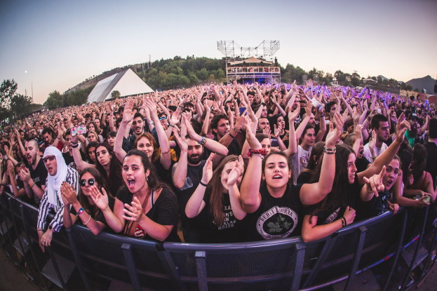 Menschenmenge auf einem Musikfestival mit erhobenen Händen vor einem Geländer, Bäume und Gebäude sind unter einem klaren blauen Himmel zu sehen.